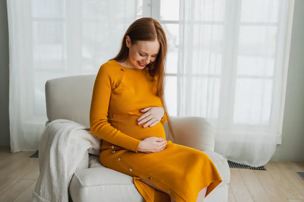 Mujer embarazada con vestido naranja, sentada cómodamente en un sillón claro, sonriendo hacia su vientre y sosteniéndolo con ambas manos. La imagen evoca ternura y cuidado, destacando el vínculo entre la madre y su bebé, así como la importancia de una buena nutrición durante el embarazo, como la ingesta de omega-3 DHA.