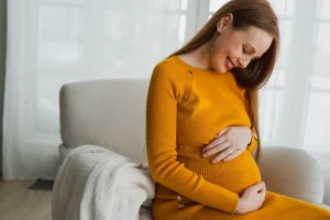 Mujer embarazada con vestido naranja sentada en un sillón claro, mirando hacia abajo y sonriendo mientras acaricia su vientre. La imagen transmite tranquilidad y alegría, reflejando la importancia de la salud materna durante el embarazo y el papel de los suplementos de omega-3 DHA para el desarrollo del bebé.